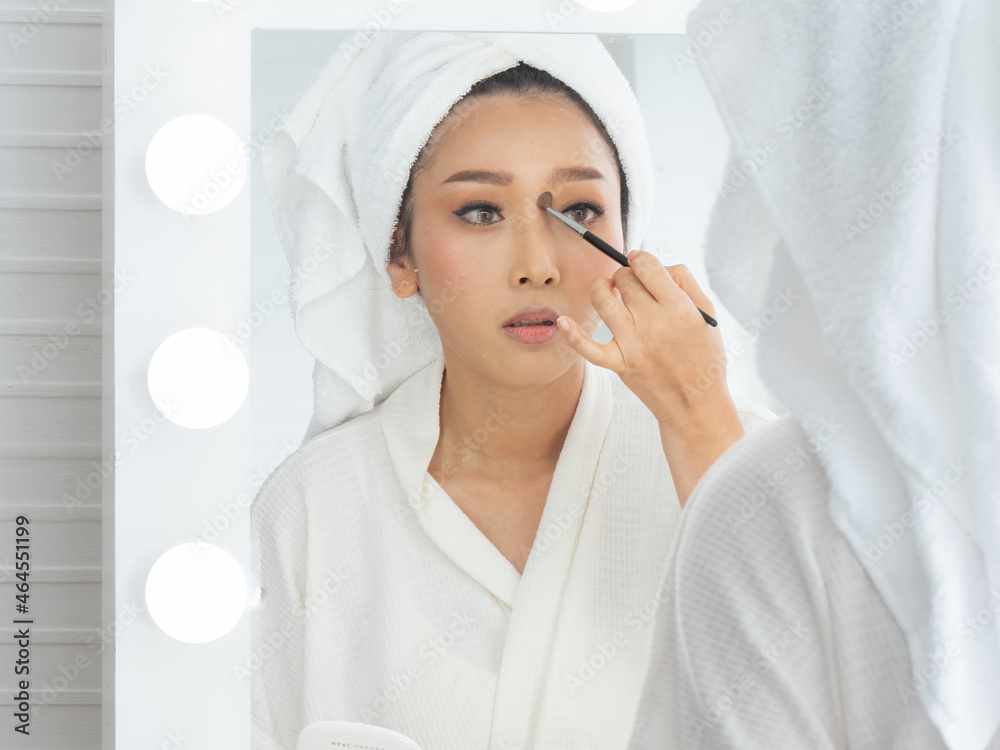 Beautiful Asian woman looking in the mirror and applying cosmetics with a small brush. Attractive woman applying her makeup reflected in a mirror with a light bulb surround at home.. Beautiful Asian woman looking in the mirror and applying cosmetics with a small brush. Attractive woman applying her makeup reflected in a mirror with a light bulb surround at home..