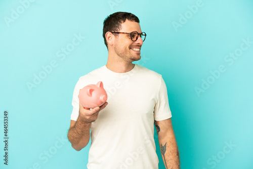 Brazilian man holding a piggybank over isolated blue background looking side