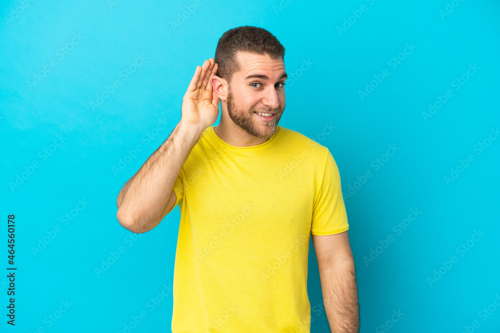 Young handsome caucasian man isolated on blue background listening to something by putting hand on the ear Young handsome caucasian man isolated on blue background listening to something by putting hand on the ear