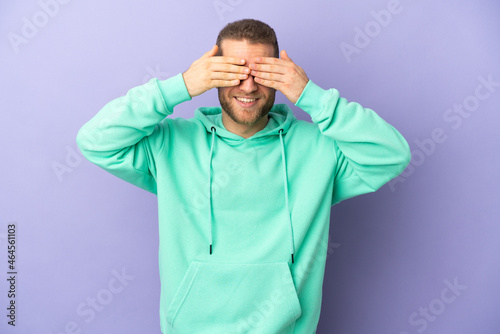 Young handsome caucasian man isolated on purple background covering eyes by hands and smiling © luismolinero