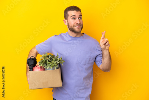 Young caucasian making a move while picking up a box full of things isolated on yellow background with fingers crossing and wishing the best