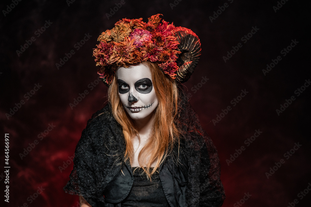 A woman in Halloween makeup and costume with horns, dried flowers, sewn-up mouth in a dark red burning room looks at the camera. All Saints' Day look Idea in calavera style A woman in Halloween makeup and costume with horns, dried flowers, sewn-up mouth in a dark red burning room looks at the camera. All Saints' Day look Idea in calavera style