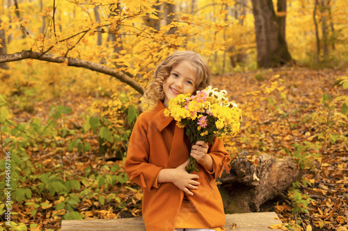 a beautiful blonde girl in a mustard-colored coat is sitting on a bench in the autumn forest with autumn flowers in her hands