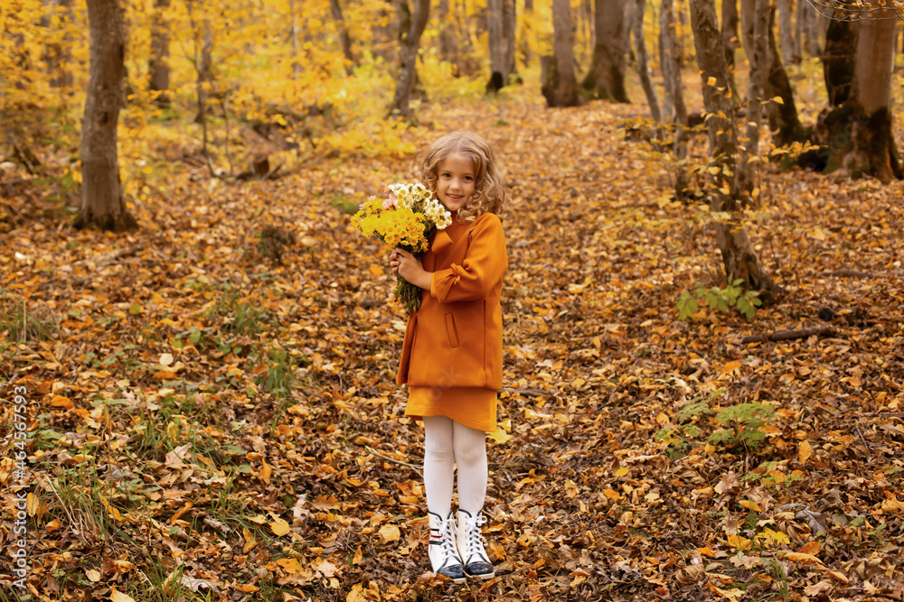 a beautiful happy smiling blonde girl in a mustard-colored coat stands in the autumn forest with autumn flowers in her hands a beautiful happy smiling blonde girl in a mustard-colored coat stands in the autumn forest with autumn flowers in her hands