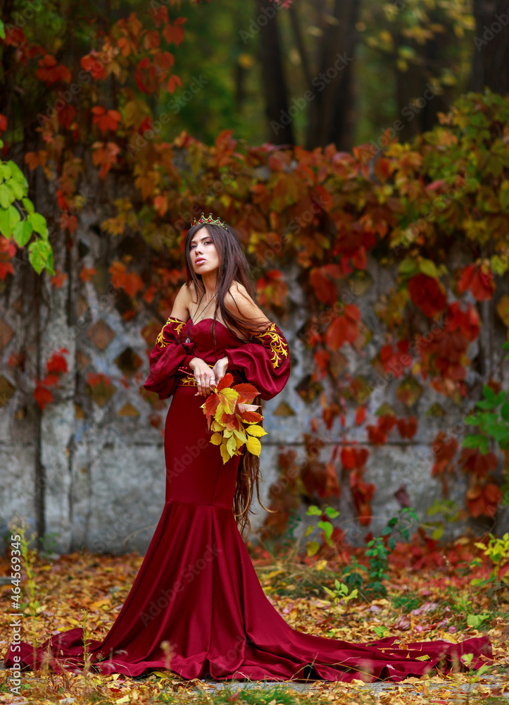 Young lady in luxurious long burgundy dress with crown on her head in colorful autumn park