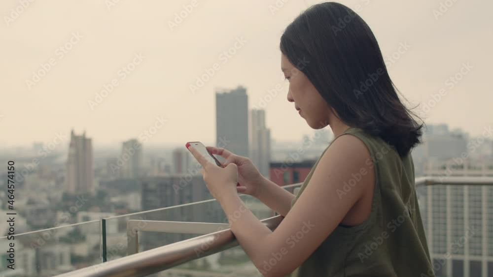 Asian female using smartphone chatting with friends while standing beside balcony on a rooftop at the condominium during sunset. Asian female using smartphone chatting with friends while standing beside balcony on a rooftop at the condominium during sunset.