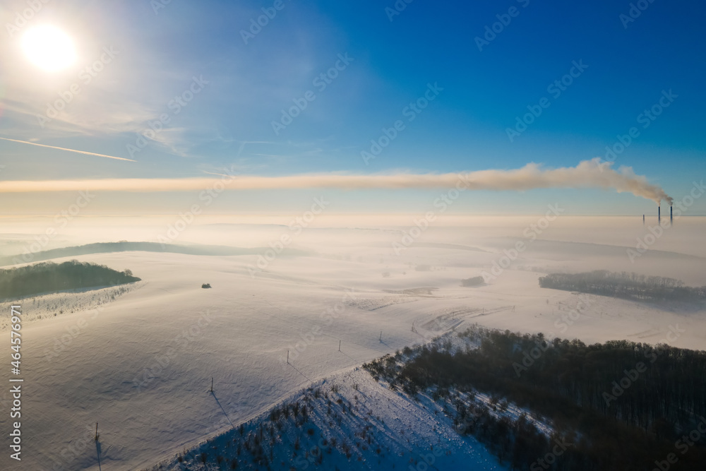 Aerial view of winter landscape with foggy countryside and distant factory pipes emmiting black dirty smoke polluting environment.