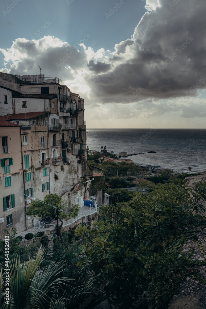 Gegenlicht-Blick kurz vor Sonnenuntergang auf die Altstadt von Tropea und das Meer mit dramatischer Wolkenkulisse, Kalabrien, Italien