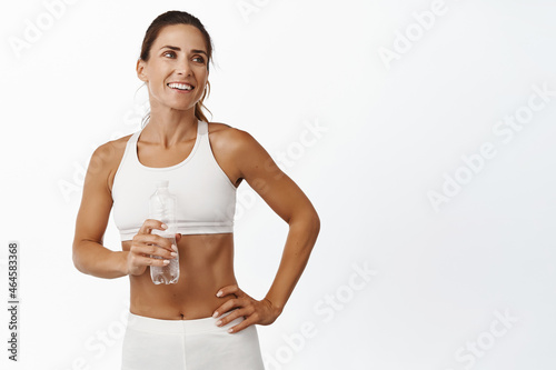 Smiling female athlete drinking water during fitness break  holding bottle and standing in activewear against white background