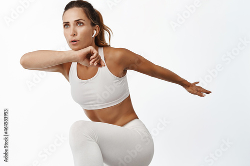 Female athlete doing fitness exercises, control breathing, listening music in wireless headphones, jumping and raising legs, white background