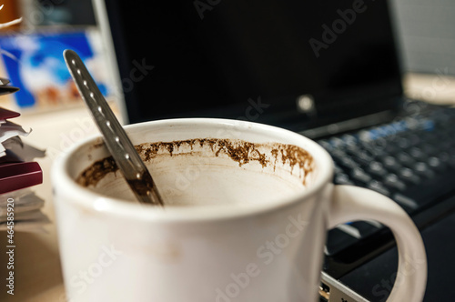 Cup of coffee with small spoon inside with open laptop lid in background - hard late working concept