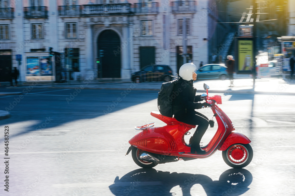 Side view of young woman with protection helmet riding fast on her red vintage scooter in central part of an old city - delivery commuting cool person