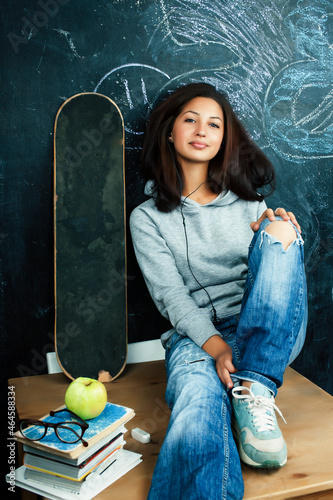 young cute teenage girl in classroom at blackboard seating on table smiling, modern hipster concept
