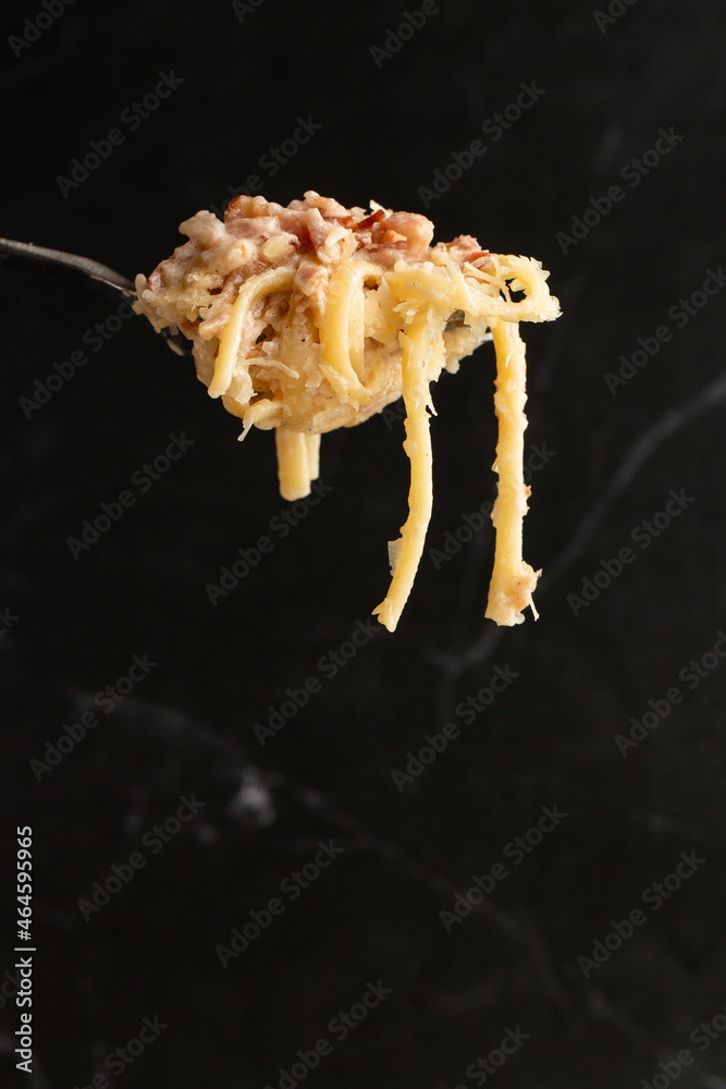 Pasta Carbonara on fork isolated. Spaghetti with bacon, parmesan, cream sauce and egg yolk on black marble background. Mediterranean gourmet food.