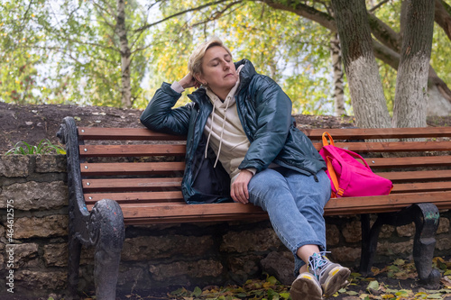 a girl in a dark jacket and with an orange backpack is sitting on a bench against the background of autumn nature, thinking and looking ahead