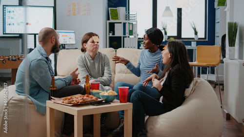 Cheerful coworkers playing guessing game with sticky notes on forehead after work for entertainment. Workmates enjoying fun activity with beer drinks, snacks and play after hours.