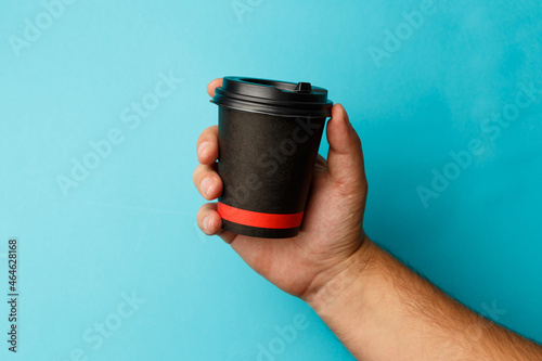 Mockup of man hand holding up a Coffee black color paper cup on blue background. Front view. Close-up of a hand. Concept coffee break. Coffee to go. Breakfast