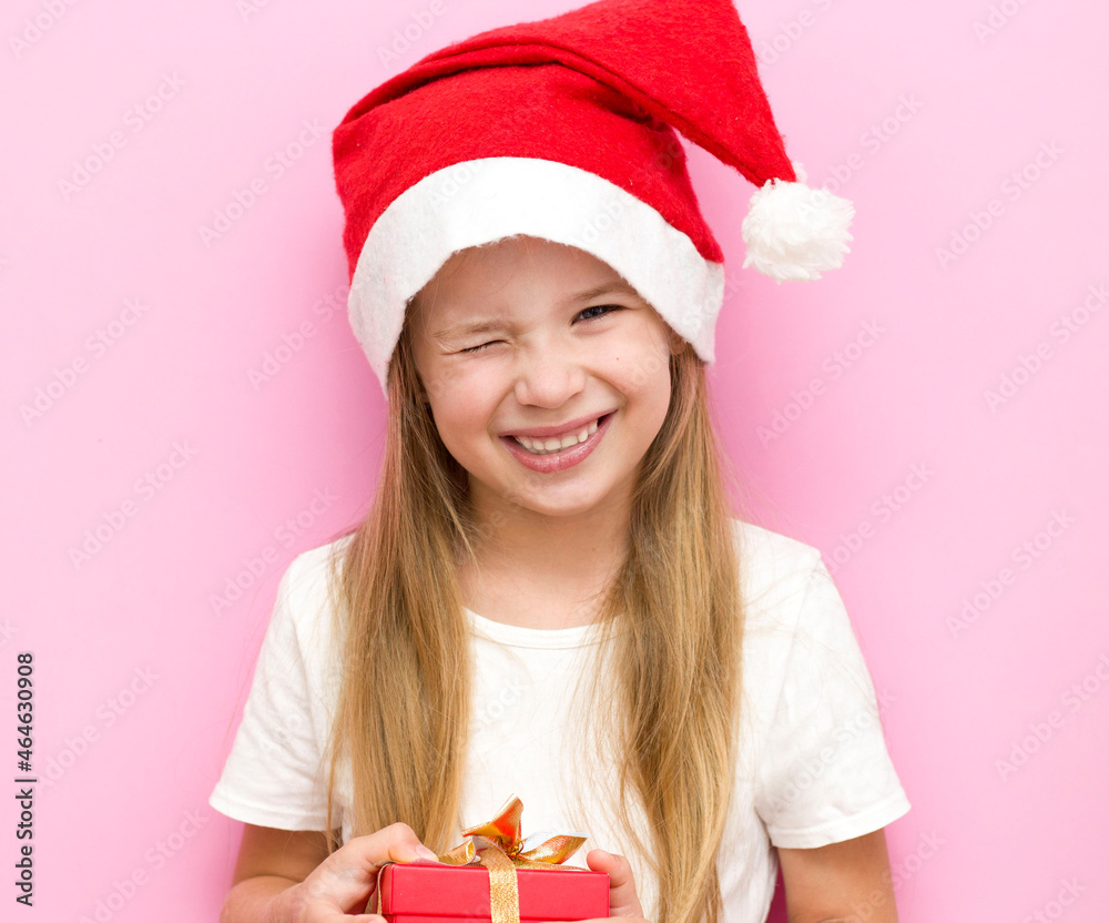 Christmas Eve! A cheerful child in a Christmas hat on a pink background with a red gift box. Christmas Eve! A cheerful child in a Christmas hat on a pink background with a red gift box.
