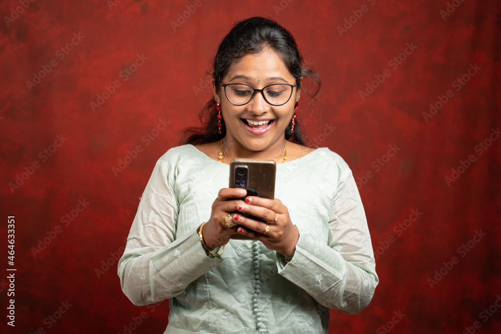 Fototapeta premium Portrait shot of surprised young Indian woman by seeing online sales and offers on mobile phone during festival - concept e-commerce discounted shopping.