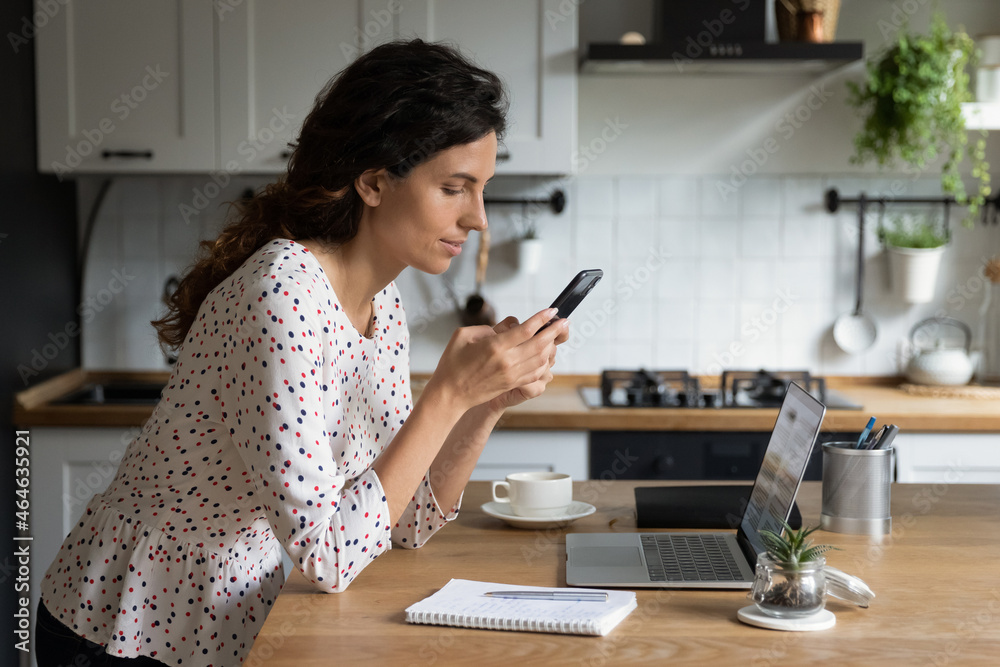 Female Hispanic remote employee working at laptop from home, using smartphone for online chatting with client, texting email. Freelancer with mobile phone making call from kitchen. Distance job Female Hispanic remote employee working at laptop from home, using smartphone for online chatting with client, texting email. Freelancer with mobile phone making call from kitchen. Distance job