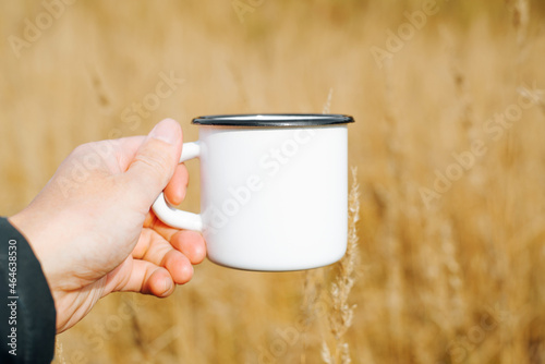 Close-up of hand showing blank white mug mockup against yellow wheat field outdoors. Enamelled travel cup with empty space for logo or branding. Camping, hike, picnic concept