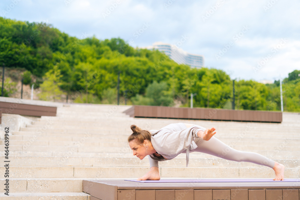 Side view of flexible young yogini woman practicing yoga exercisers on fit mat in summer day outdoors in city park. Calm female with beautiful body in sportswear performing asana pose outside.