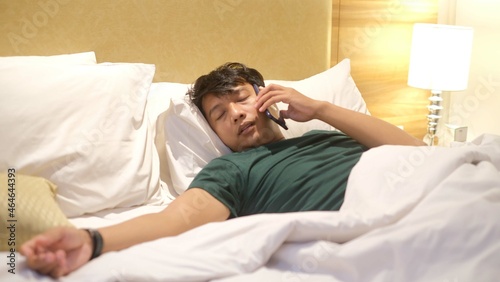 Sleepy Asian man is picking up the phone on his comfortable bed. Boring freelancer lifestyle concept.