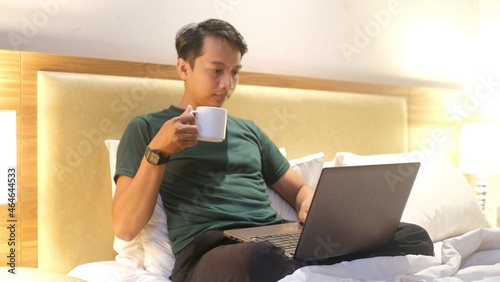 Happy creative Asian man working with laptop while holding cup on mattress