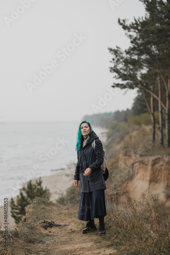 beautiful woman with blue hair in a raincoat and with a backpack stands on a hill above the beach
