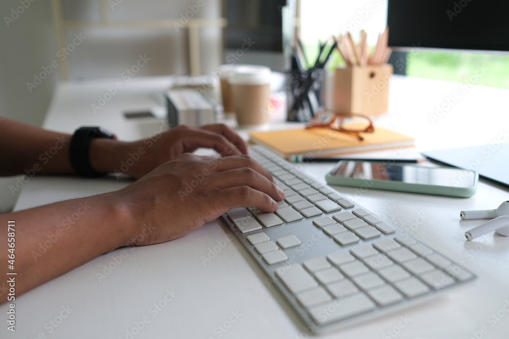 Fototapeta premium Cropped image of programmer hand typing on a keyboard at the white working desk that surrounded by a smartphone, wireless earphone and various equipment.