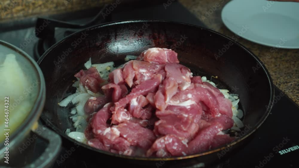 Pieces of pork flesh with chopped onions during frying in an old frying pan, top view. A woman's hand turns it over with a spoon and mixes it so that it doesn't burn. Potatoes are being cooked nearby. Pieces of pork flesh with chopped onions during frying in an old frying pan, top view. A woman's hand turns it over with a spoon and mixes it so that it doesn't burn. Potatoes are being cooked nearby.