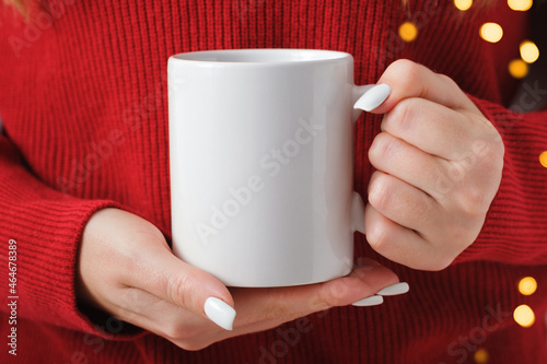 Female hands hold a mockup white tea mug in a red winter sweater, gold bokeh