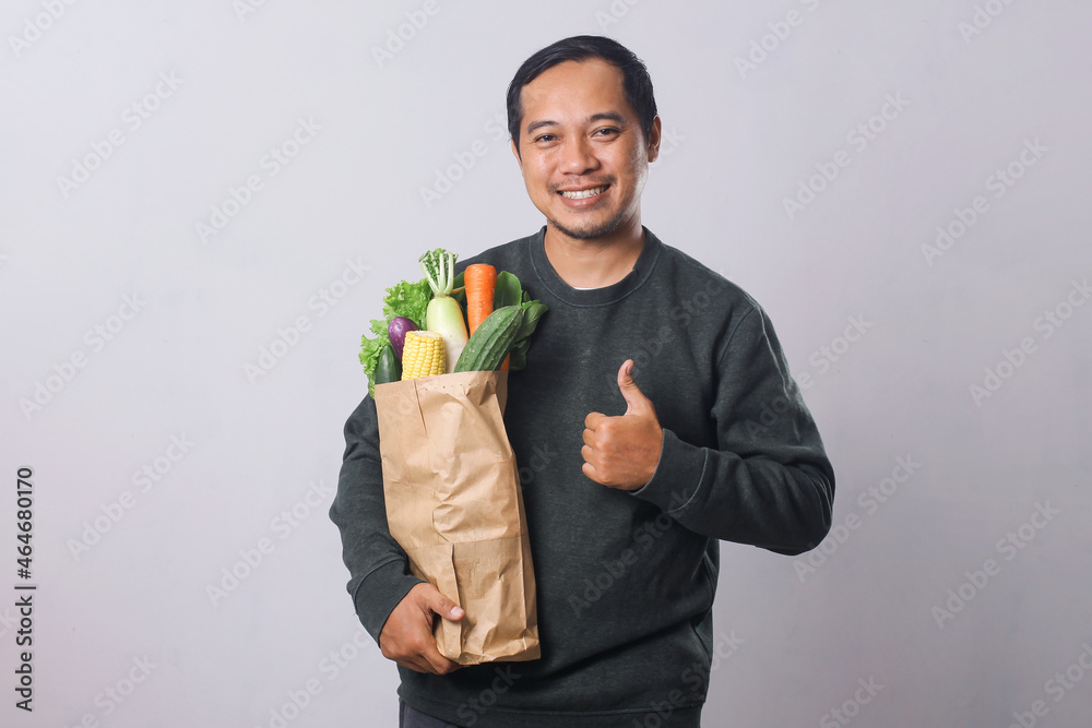 Asian man holding grocery shopping bag with vegetables with thumbs up gesture for healthy lifestyle concept Asian man holding grocery shopping bag with vegetables with thumbs up gesture for healthy lifestyle concept
