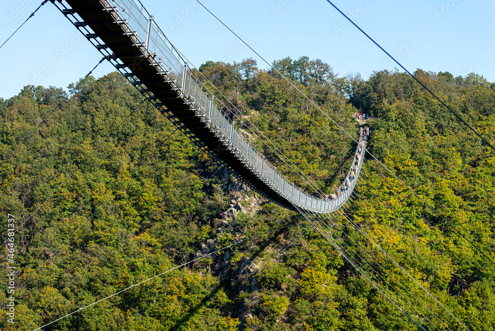 A hanging wooden bridge with steel ropes seen from the side against a blue sky, visible tourists on the bridge.