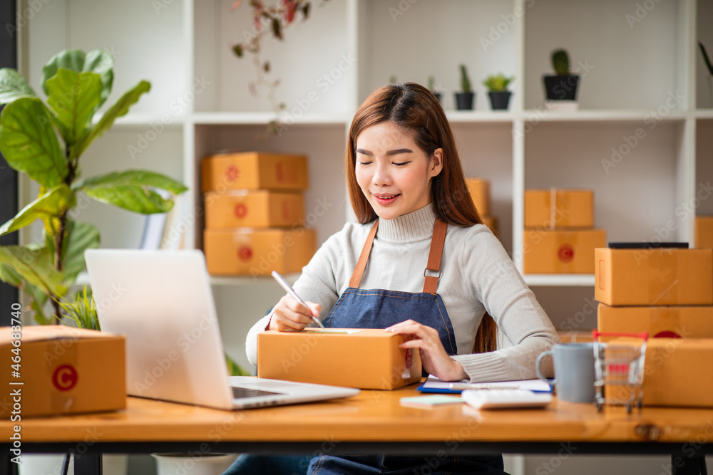 Stylish small business owner Young Asian woman using laptop computer in her home office, small business entrepreneur SME, working with a box at home, Online selling, e-commerce, packing concept