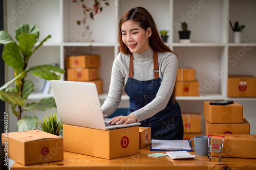 Stylish small business owner Young Asian woman using laptop computer in her home office, small business entrepreneur SME, working with a box at home, Online selling, e-commerce, packing concept © David
