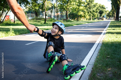 The boy is rollerblading on the track in the city park. The child learns to roller. An active walk with a child on the street. 
Dad helps the child by the hand to get up from the asphalt after a fall.