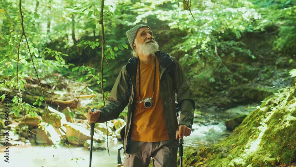 Happy Elderly Man with a Backpack and Nordic Walking Sticks Standing Near the River Enjoying Nature While Hiking in the Forest. Active Retirement, Tourism and Hobby Concept. Happy Elderly Man with a Backpack and Nordic Walking Sticks Standing Near the River Enjoying Nature While Hiking in the Forest. Active Retirement, Tourism and Hobby Concept.