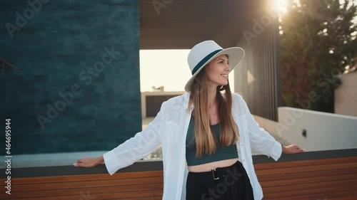 Beautiful young woman in stylish summer clothes and hat leaning on wooden fence whike posing on city street. Caucasian female smiling on camera and enjoying sunny weather outdoors. photo