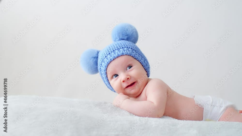 Happy newborn baby in a blue hat with pompoms looks into the camera. Smiling two-month-old infant is lying on a blanket. Beautiful, happy little child with blue eyes. Cheerful White kid. SLOW MOTION Happy newborn baby in a blue hat with pompoms looks into the camera. Smiling two-month-old infant is lying on a blanket. Beautiful, happy little child with blue eyes. Cheerful White kid. SLOW MOTION