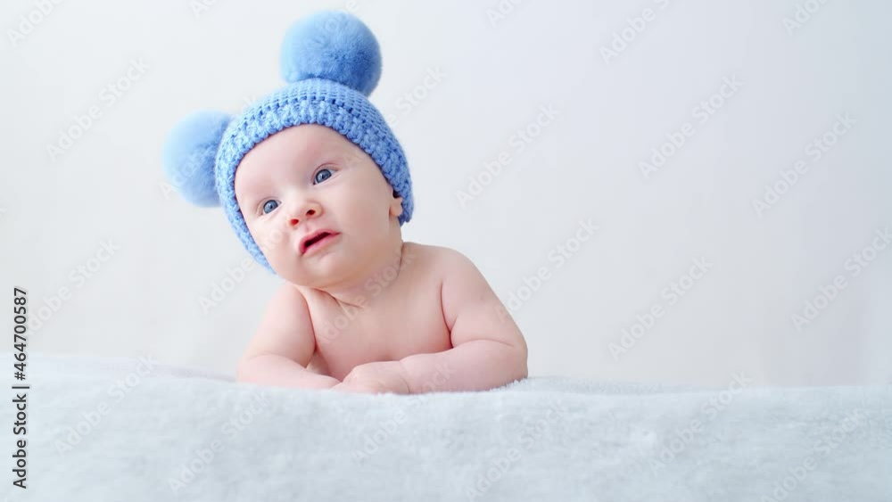 Fun Beautiful, cute little infant with blue eyes looks into camera. SLOW MOTION. Sweet newborn baby in a blue hat with pompoms looks into the camera. Calm two-month-old kid is lying on a blanket. Fun Beautiful, cute little infant with blue eyes looks into camera. SLOW MOTION. Sweet newborn baby in a blue hat with pompoms looks into the camera. Calm two-month-old kid is lying on a blanket.