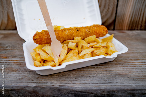 Close and selective focus on a polystyrene carton comprising a battered jumbo sausage and traditional chip shop chips cooked in beef dripping on an outdoor wooden table