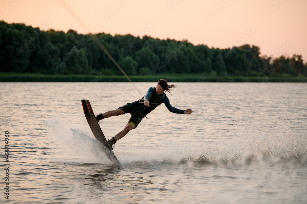 Handsome guy holds rope and energetically riding wakeboard on splashing river water.