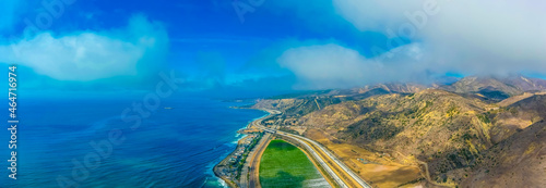 a breathtaking aerial panoramic shot of the coastline with vast blue ocean water, lush green farmland and cars on the roads along the beach with blue sky and clouds at Rincon Beach in Ventura County photo