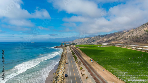 a breathtaking aerial shot of the vast blue ocean water, lush green farmland, cars on the road, majestic mountain ranges, thick cloud cover with blue sky at Rincon Beach in Ventura County California 