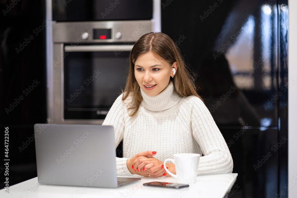 Fototapeta premium Young woman sitting at home in the kitchen and talking on video communication via laptop