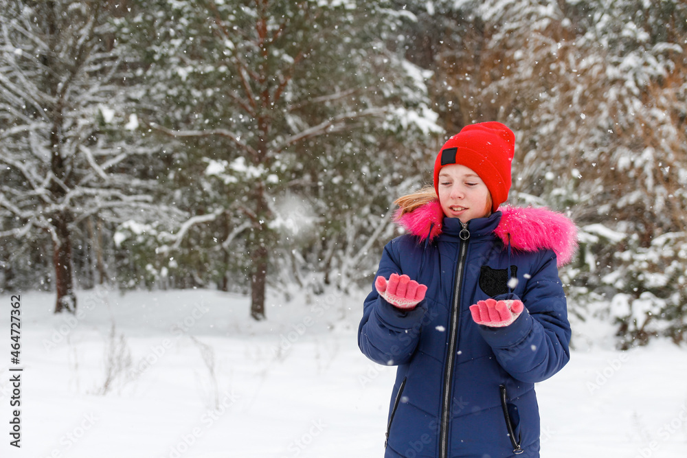 Fototapeta premium Portrait of preteen happy girl in red warm hat catching snowflakes outside on nature winter snowy forest background. Pretty child outdoor, cold weather. Caucasian kid. Teenage riot. Winter joy