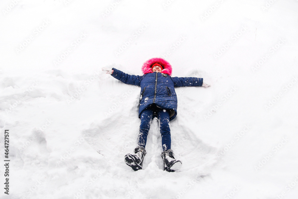 Preteen smiling girl in bright pink warm hood making snow angel while lying on snow outside background. Playful child outdoor, cold weather. Caucasian kid. Fun holidays and vacations. Winter enjoy