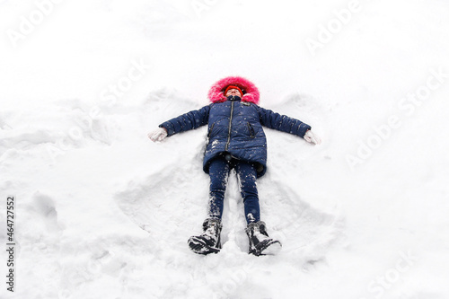 Preteen happy girl in bright pink warm hood making snow angel while lying on snow outside background. Pretty child outdoor, cold weather. Caucasian kid. Fun holidays and vacations. Winter enjoy
