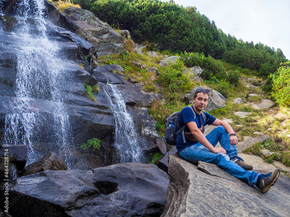 Tourist rises in the mountains to the highest waterfall of Ukraine under the mountain Goverla. Man sits on the background with waterfall of wild mountain slopes of the Carpathian Mountains. 